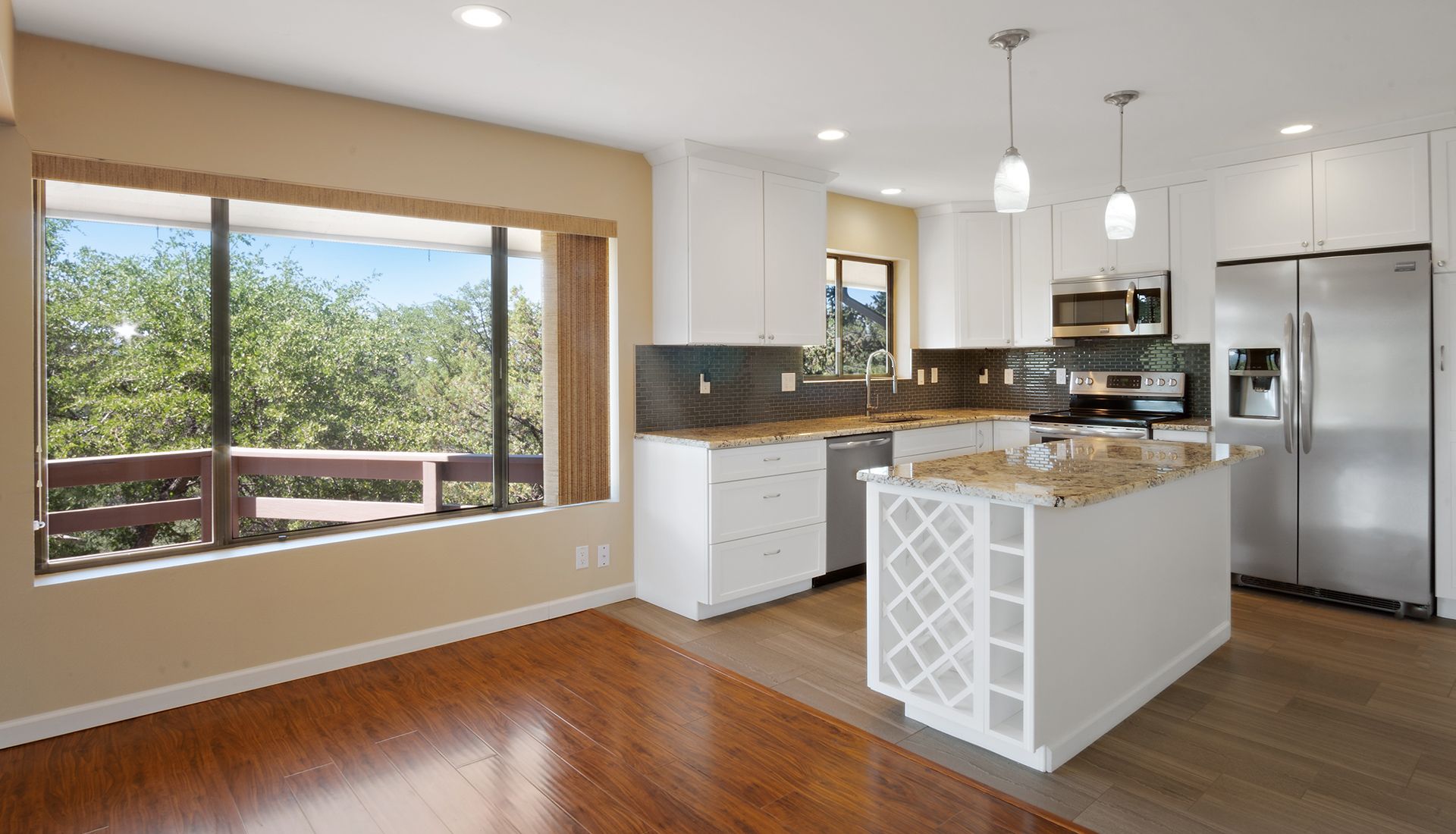 Modern kitchen with white cabinetry, stainless steel appliances, granite island, and a large window overlooking trees.