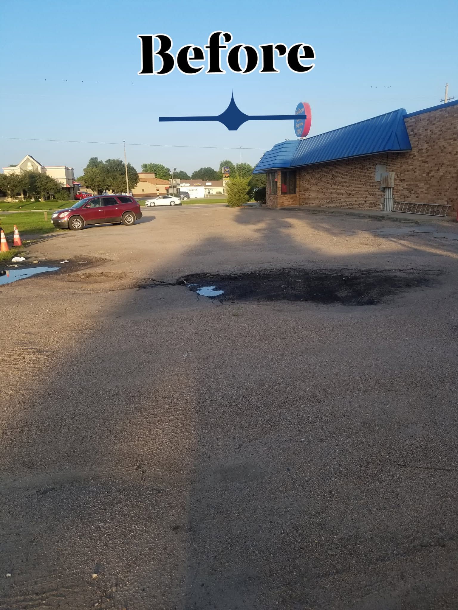 Before photo of a gravel parking lot beside a brick building with a blue roof, marked by a dark pavement stain.