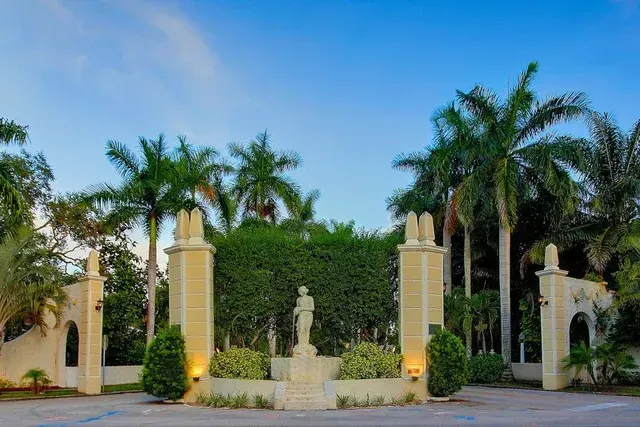 A statue of a man stands in front of a gated entrance surrounded by palm trees.