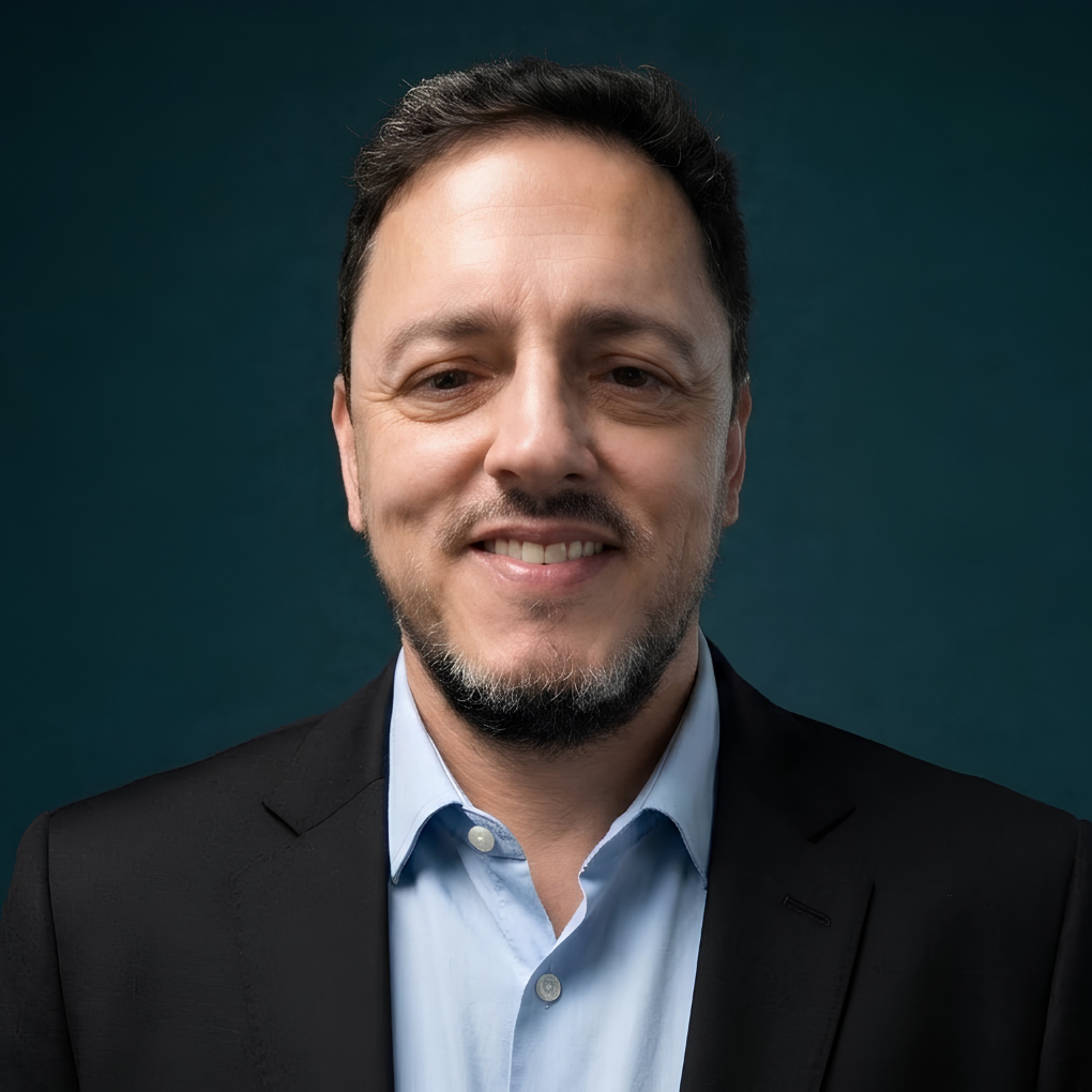 Retrato de um homem sorridente com blazer escuro e camisa azul-clara contra um fundo verde-azulado.