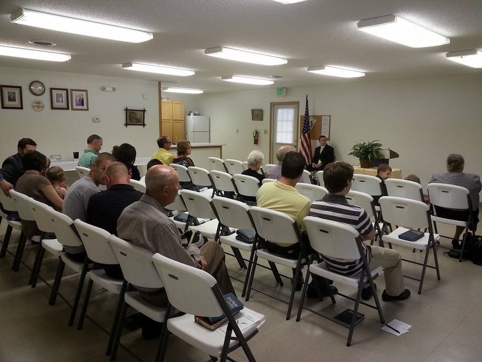 People seated in folding chairs in a small meeting room with fluorescent lights and wall art.