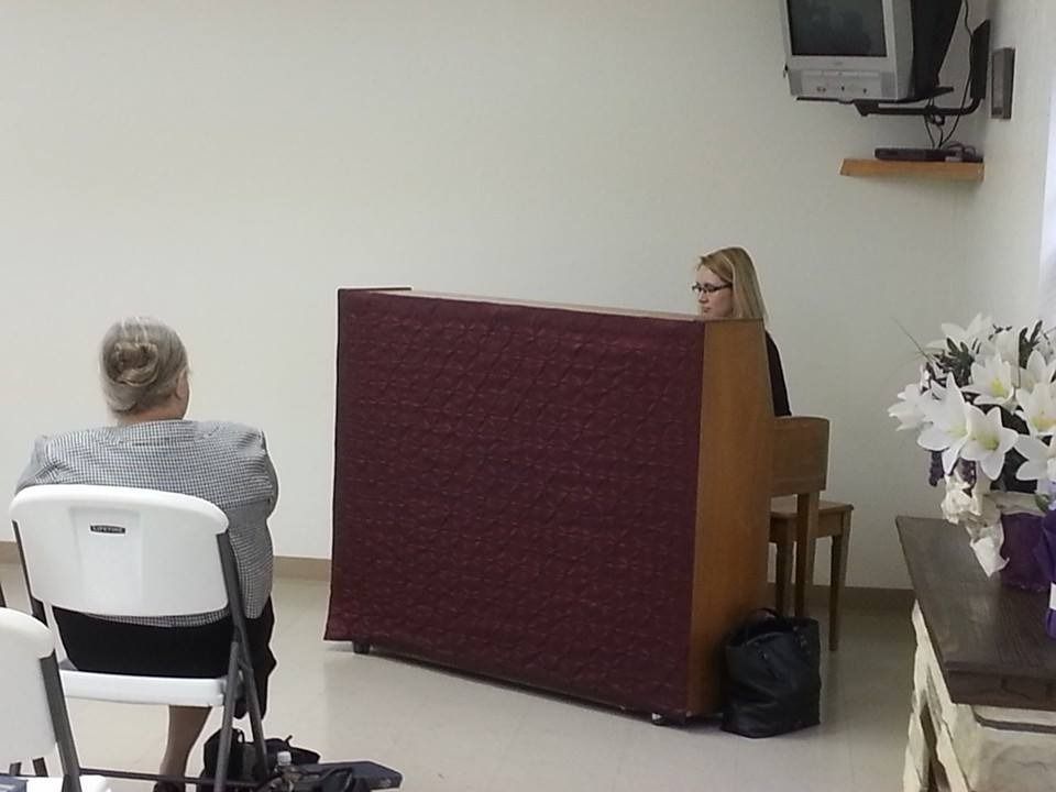 Woman seated behind a red panel in a small room, with a man seated nearby and a TV on the wall.