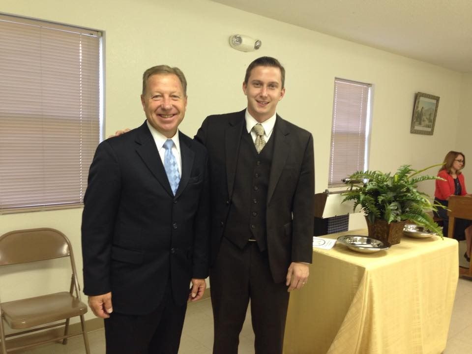 Two men in suits standing indoors beside a table with a plant and yellow tablecloth