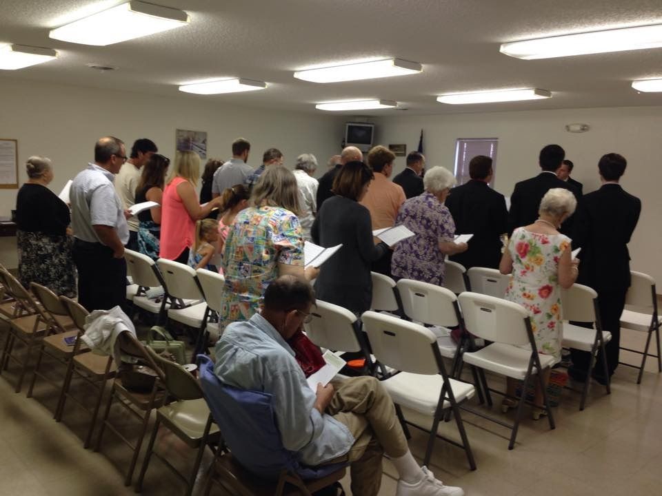 People standing in a small community meeting room with rows of folding chairs and fluorescent lighting