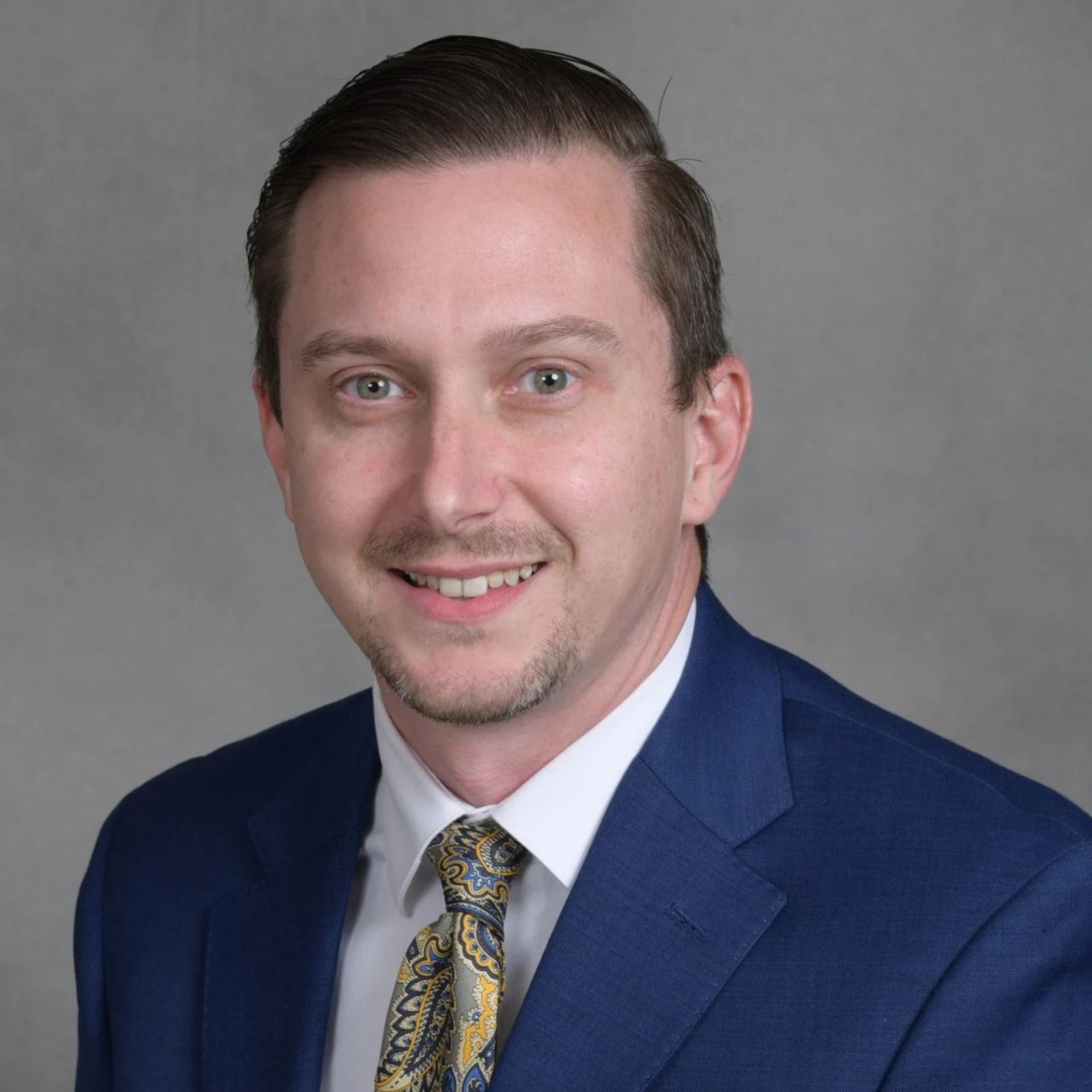 Headshot of a man in a blue suit and patterned tie on a gray background