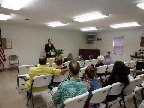 Community meeting in a plain room, with a speaker at the front and seated audience members.