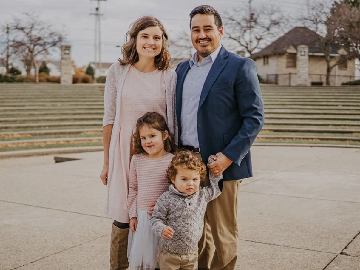 Family of four standing outdoors on concrete steps, with houses and a cloudy sky in the background.