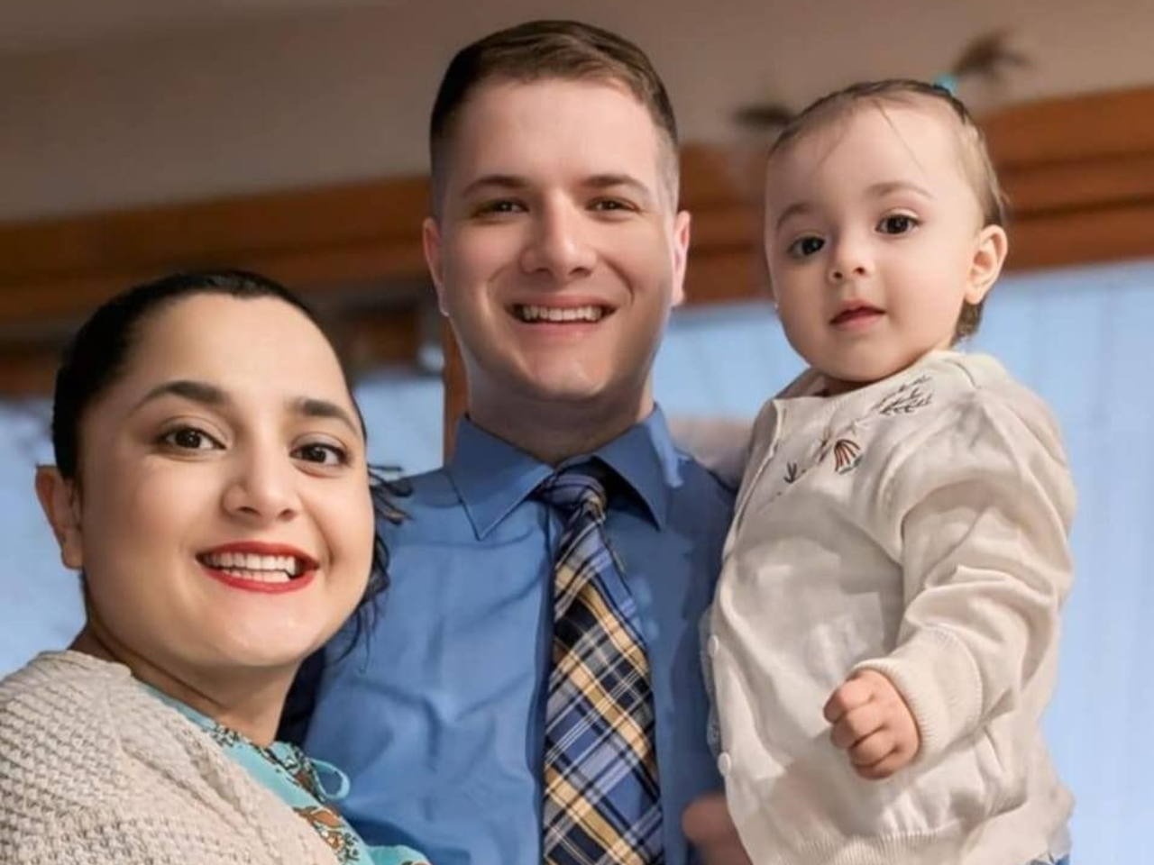 Three smiling people posing indoors, one holding a baby, in front of a warm-toned background