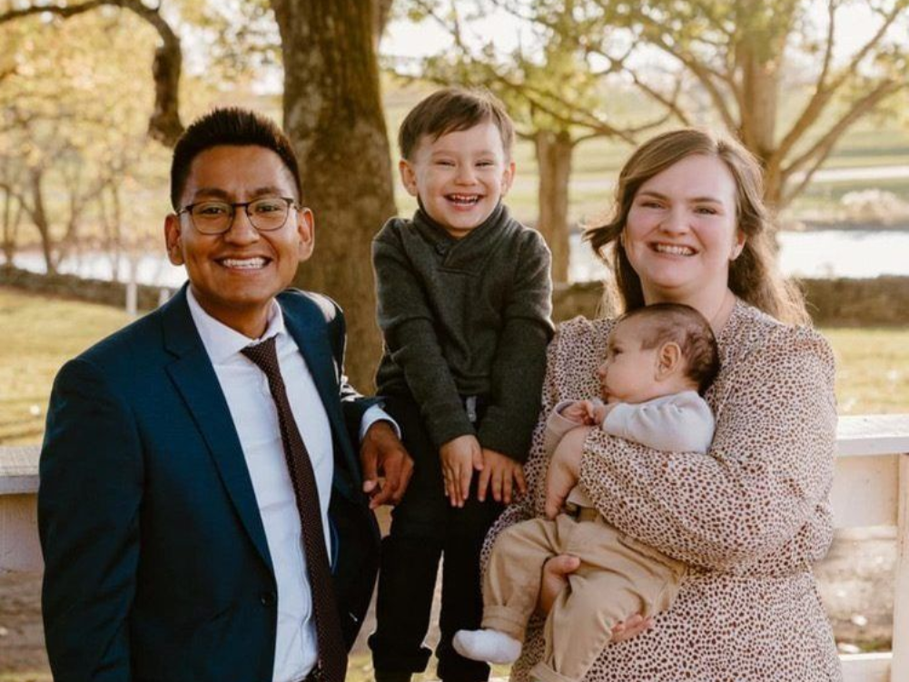 Family of four posing outdoors, with one child on a man's shoulders and a baby in a woman's arms.