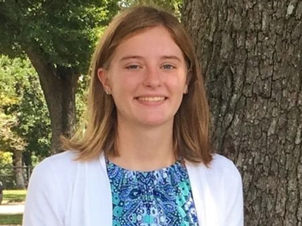 Smiling person in a blue patterned top and white cardigan standing in front of a tree trunk outdoors