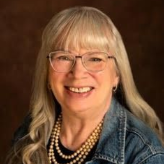 Smiling woman with glasses and pearl necklace, wearing a denim jacket against a dark backdrop