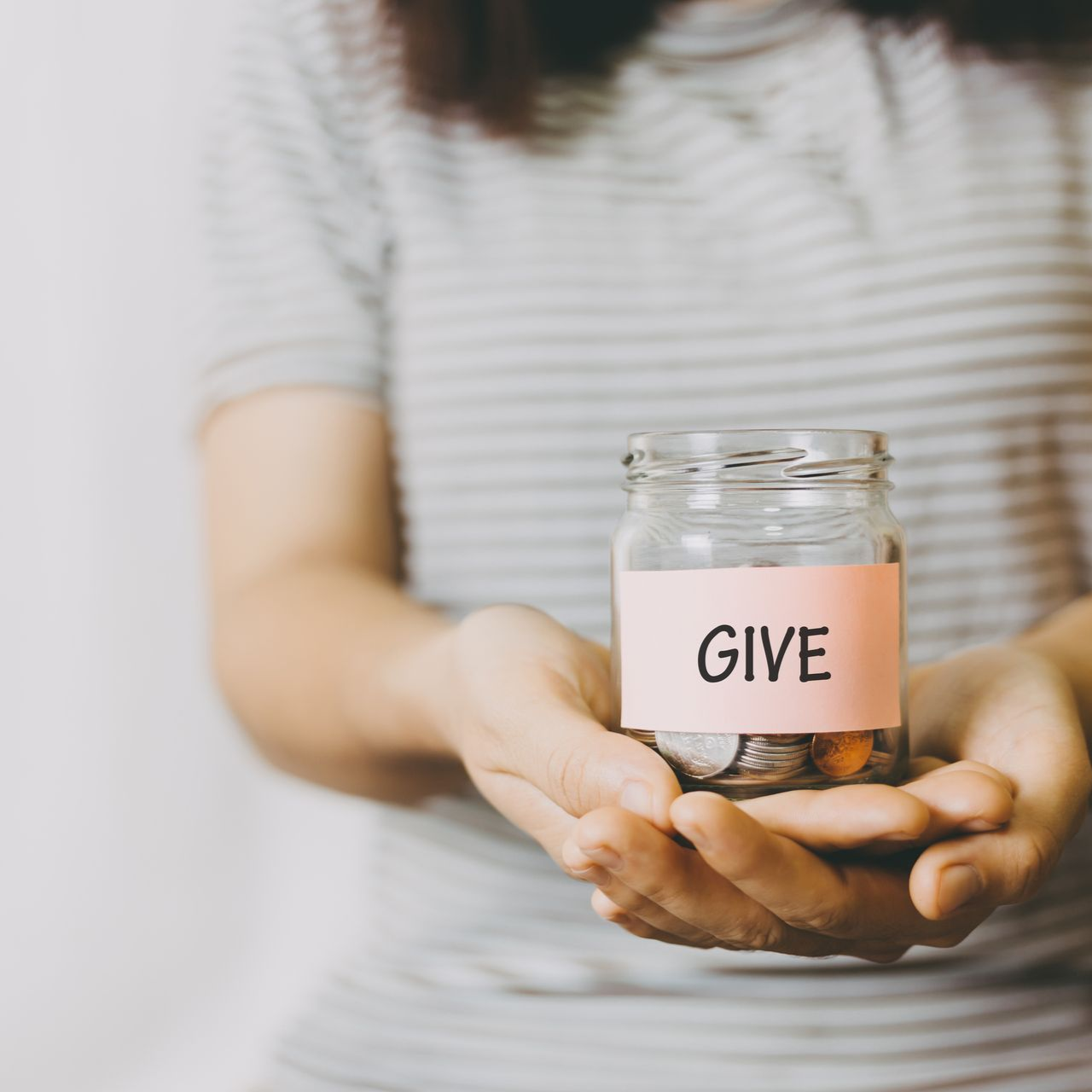 Hands holding a glass jar labeled “GIVE” with coins inside, on a soft striped background