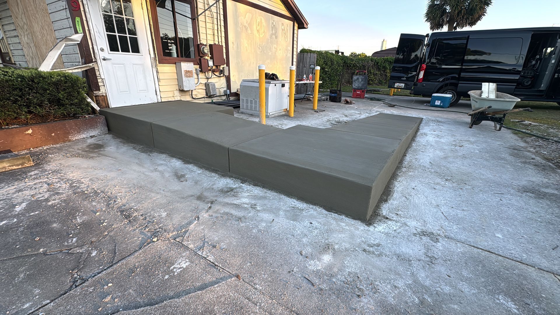 Freshly poured concrete patio beside a house, with yellow posts and outdoor furniture nearby.