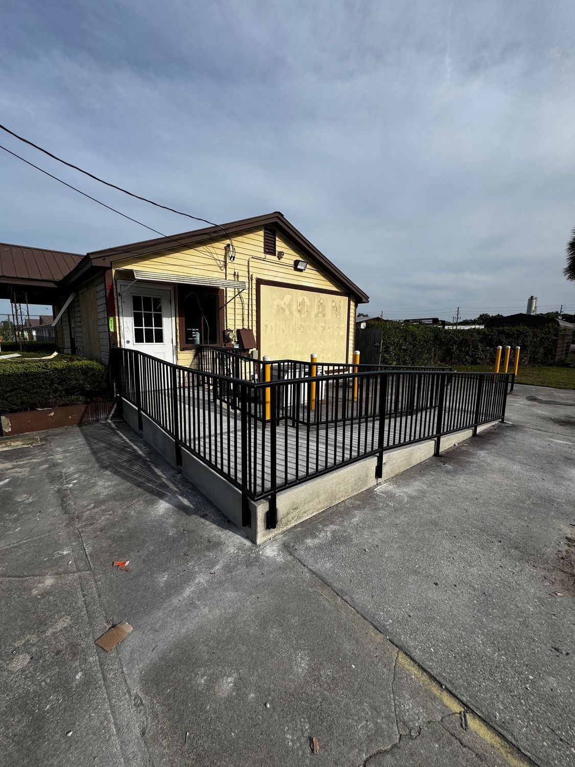 Small yellow building with a black railing on a paved lot under a cloudy sky