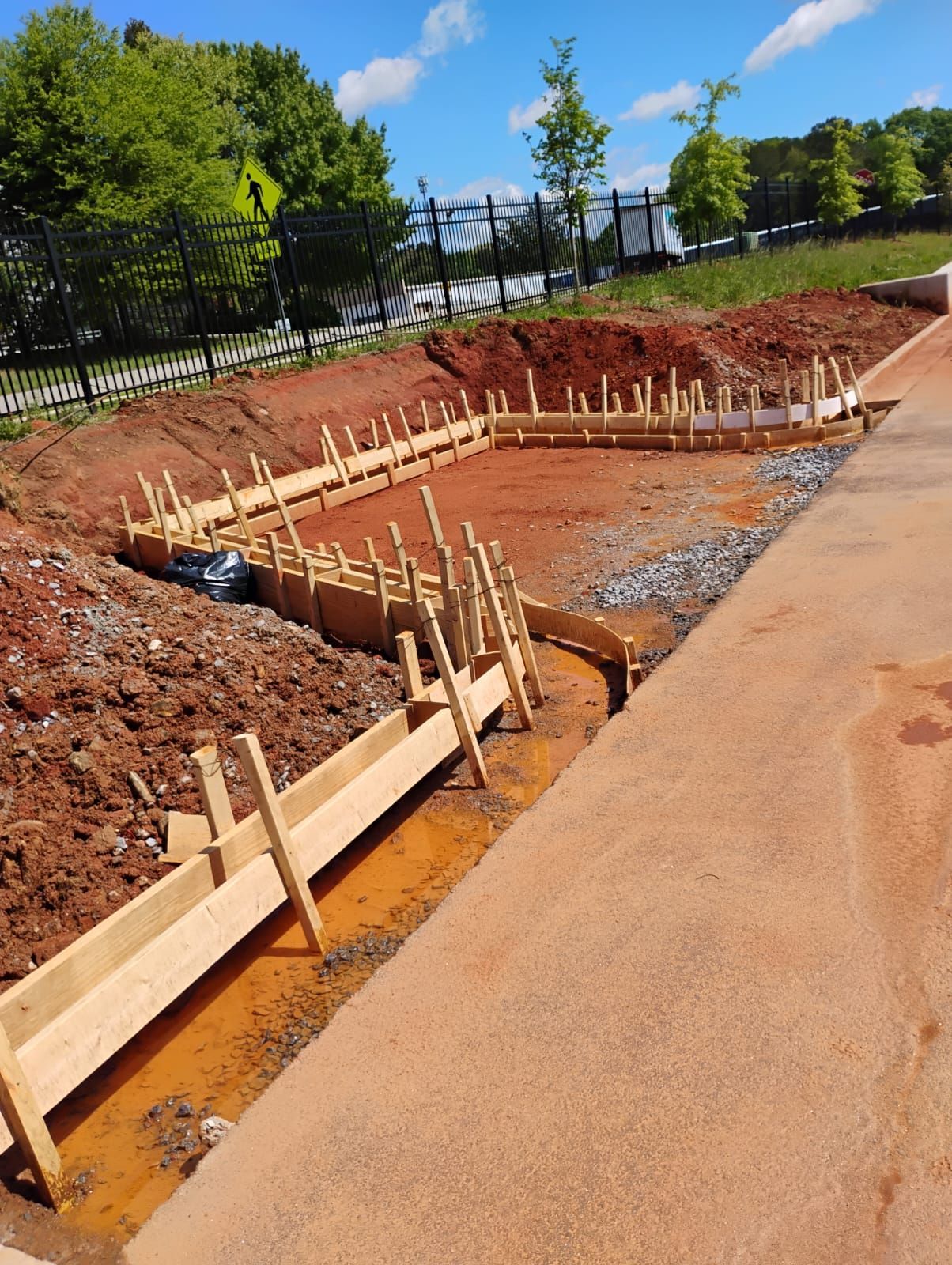 Construction site with a concrete foundation excavation, wooden forms, and orange dirt beside a road