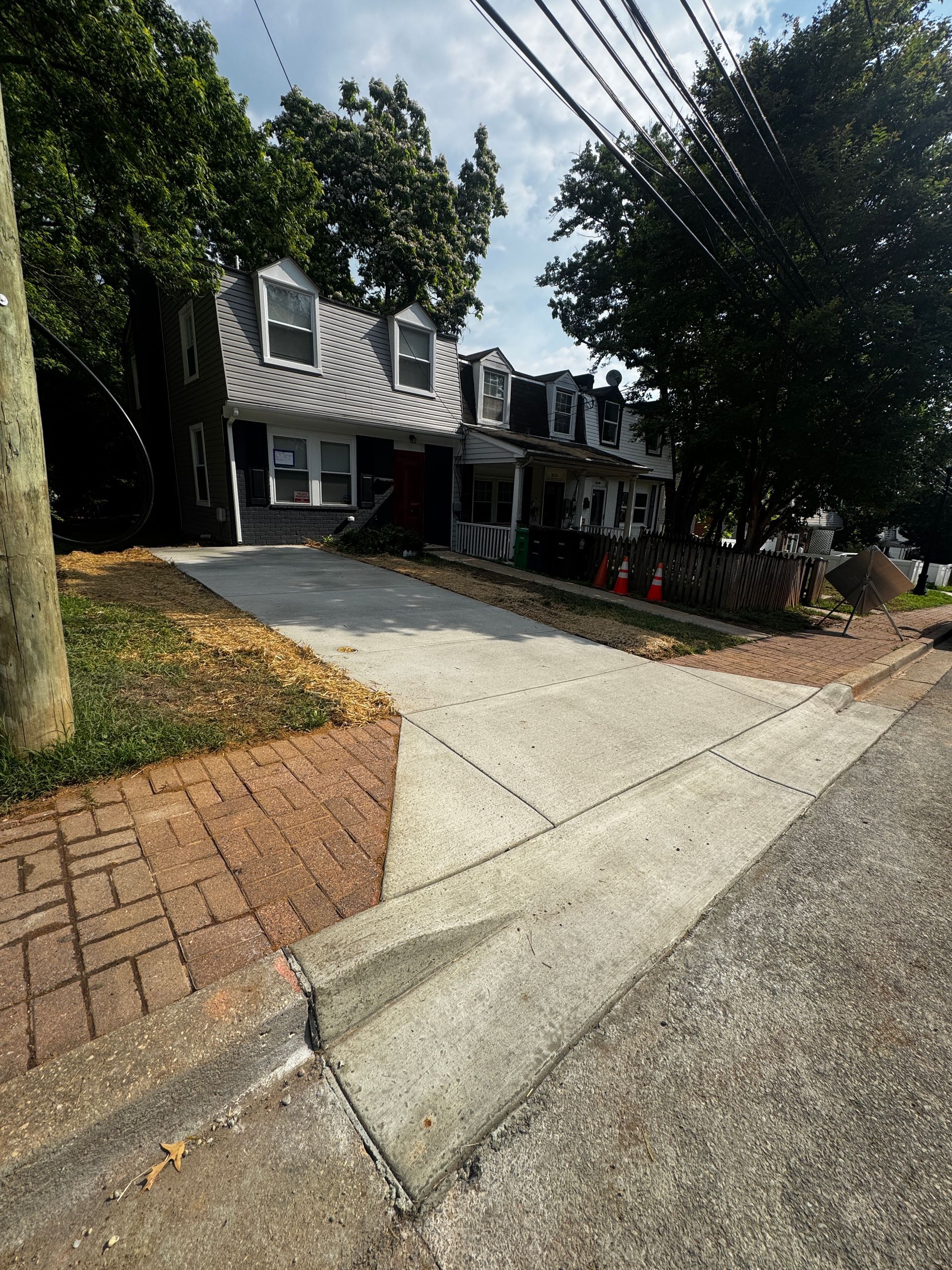 Brick driveway leading to a gray house with white trim, trees overhead, and a landscaped front yard