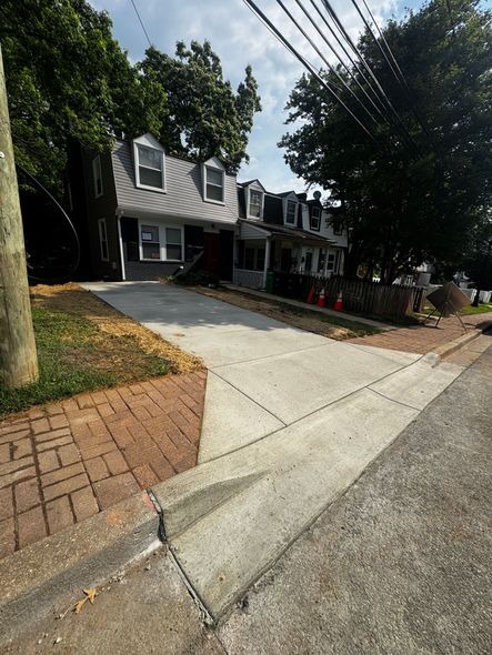 Brick driveway leading to a gray house with white trim, trees overhead, and a landscaped front yard