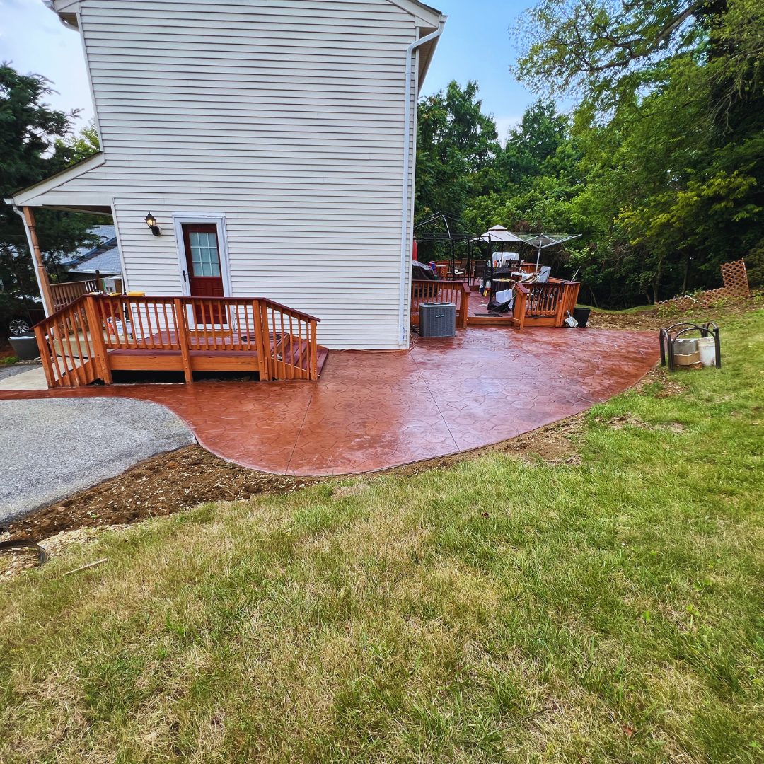 White house with a wooden deck and red concrete patio in a green backyard.