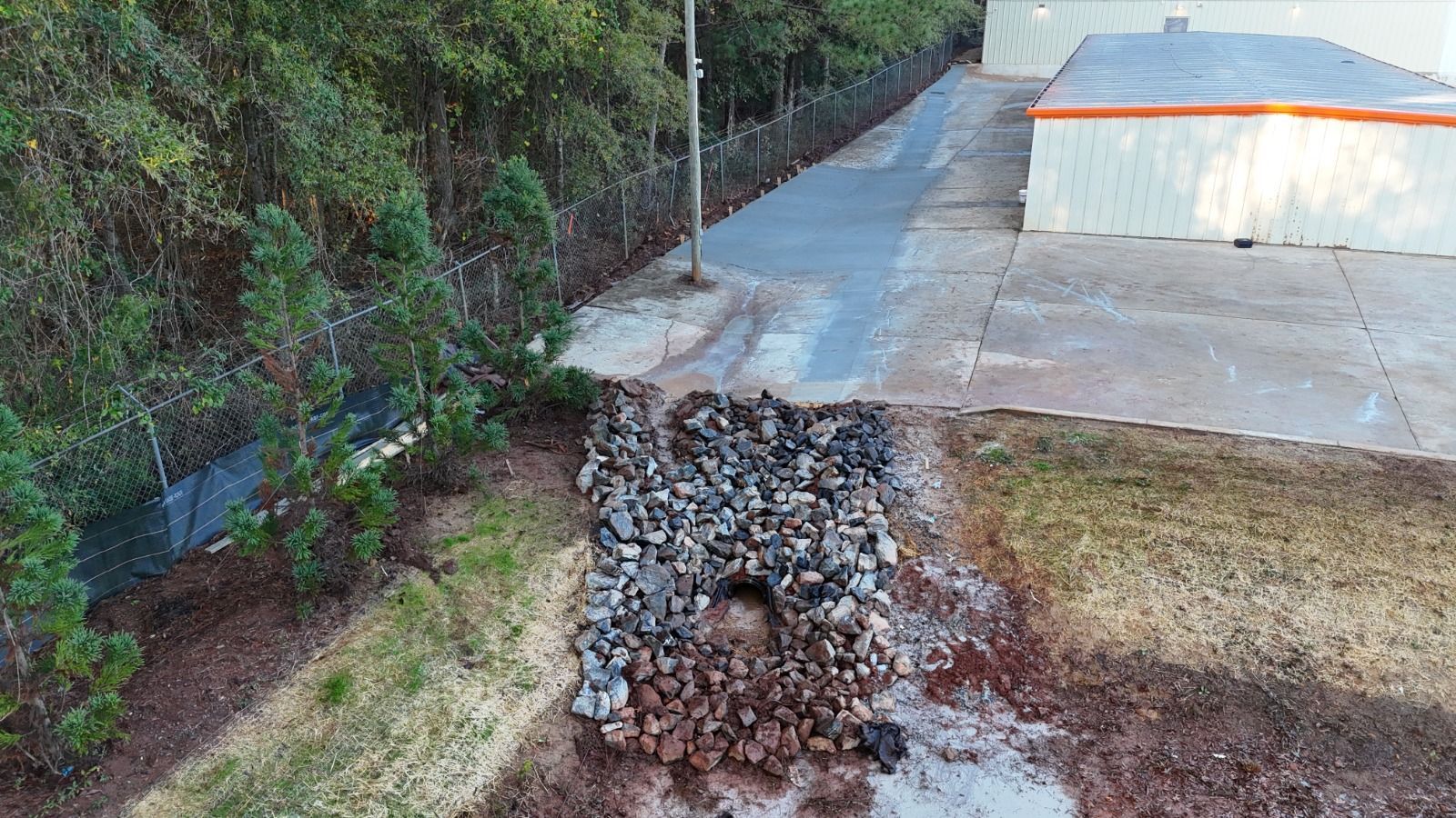 A rocky drainage channel beside a concrete path and building, with trees and grass on both sides.
