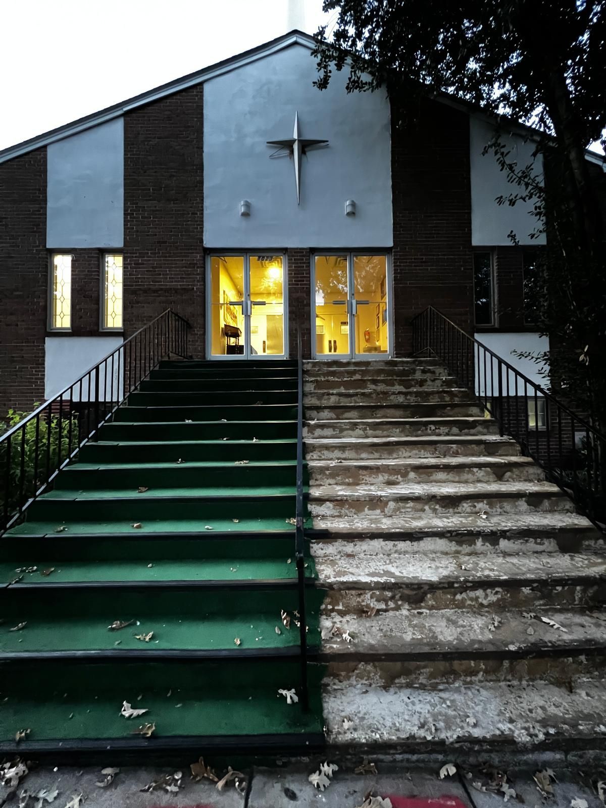 Front view of a church entrance with green and concrete stairs, lit doors, and a cross above the doorway