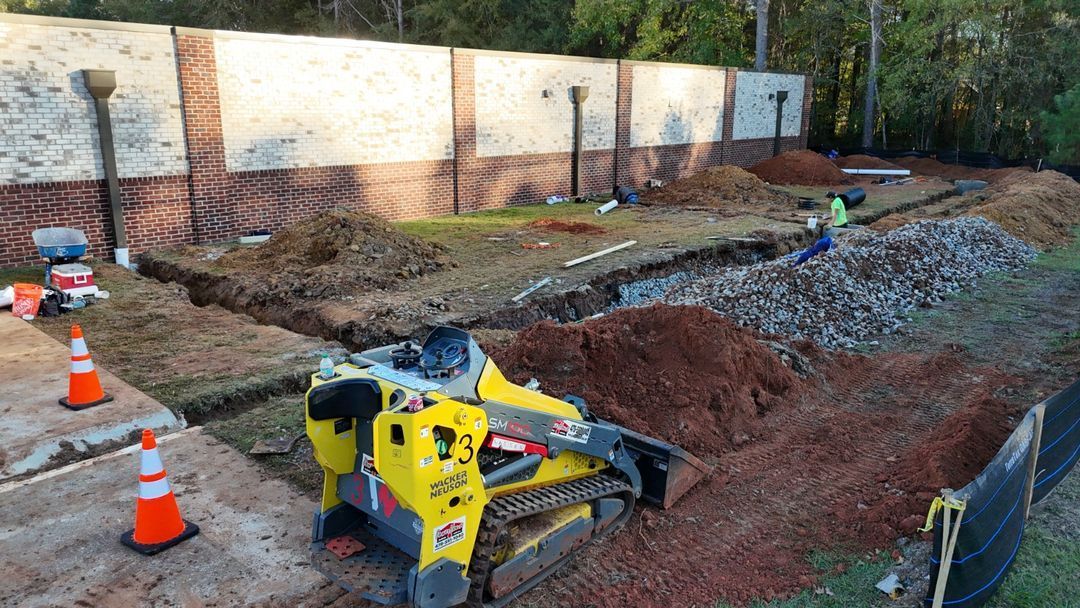 Construction site with yellow excavator by a trench, gravel pile, and orange traffic cones along a fence.
