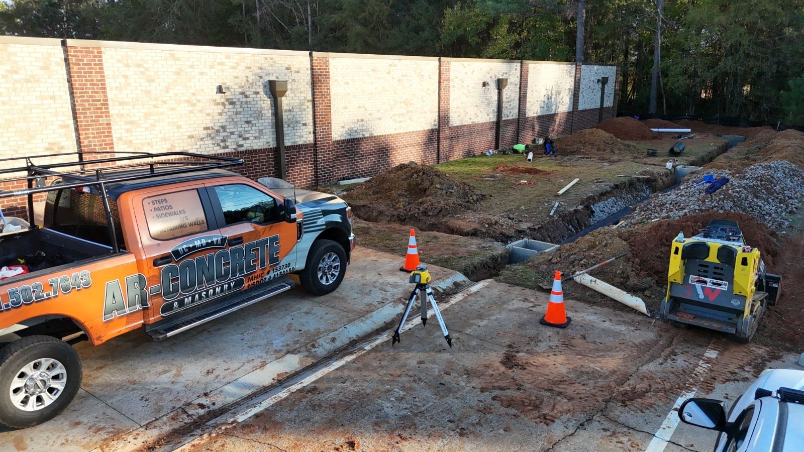 Construction site with orange utility truck, cones, and a compact excavator beside a trench and brick wall
