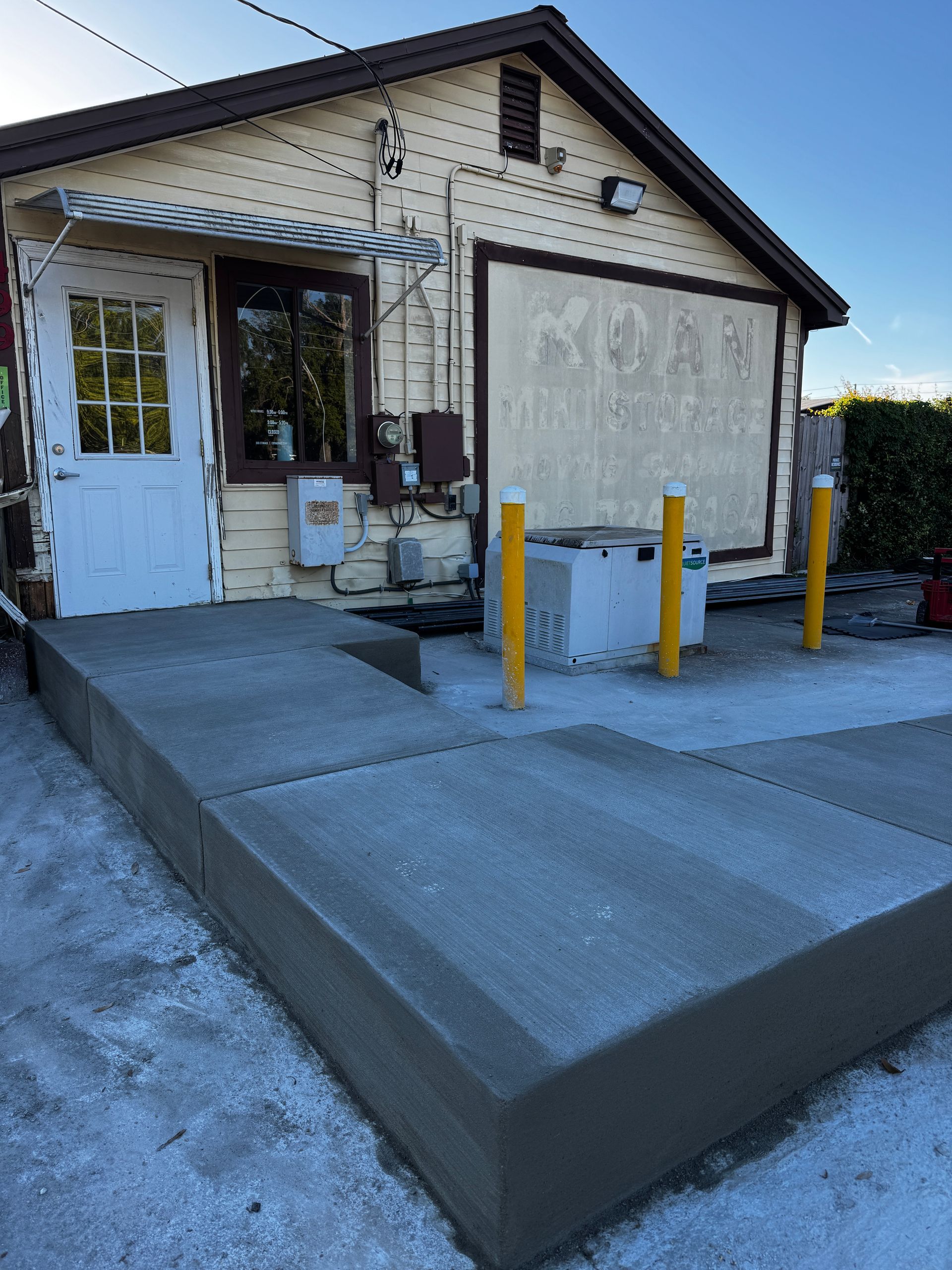Small brick building entrance with a concrete ramp, blue door, and yellow bollards outside.