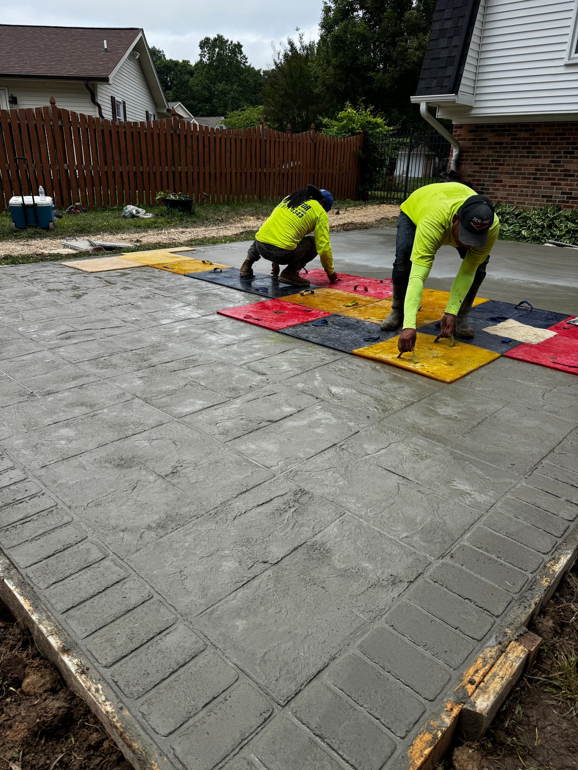 Two workers in neon jackets install colorful pavers on a gray concrete driveway outdoors.