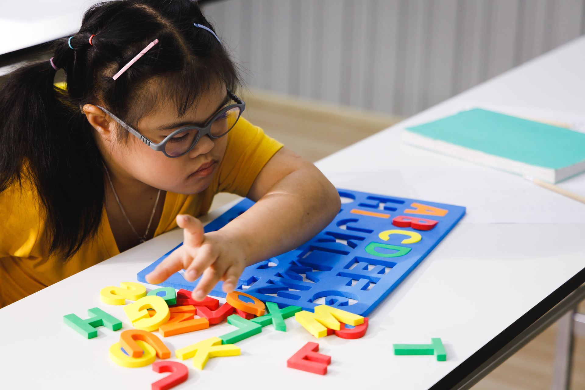 Girl with Down syndrome wearing glasses plays with colorful alphabet letters at a table.