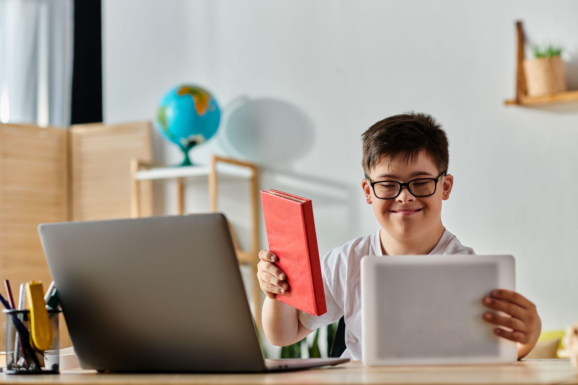 Smiling boy with Down syndrome holding a red book and tablet at his desk for special ed.