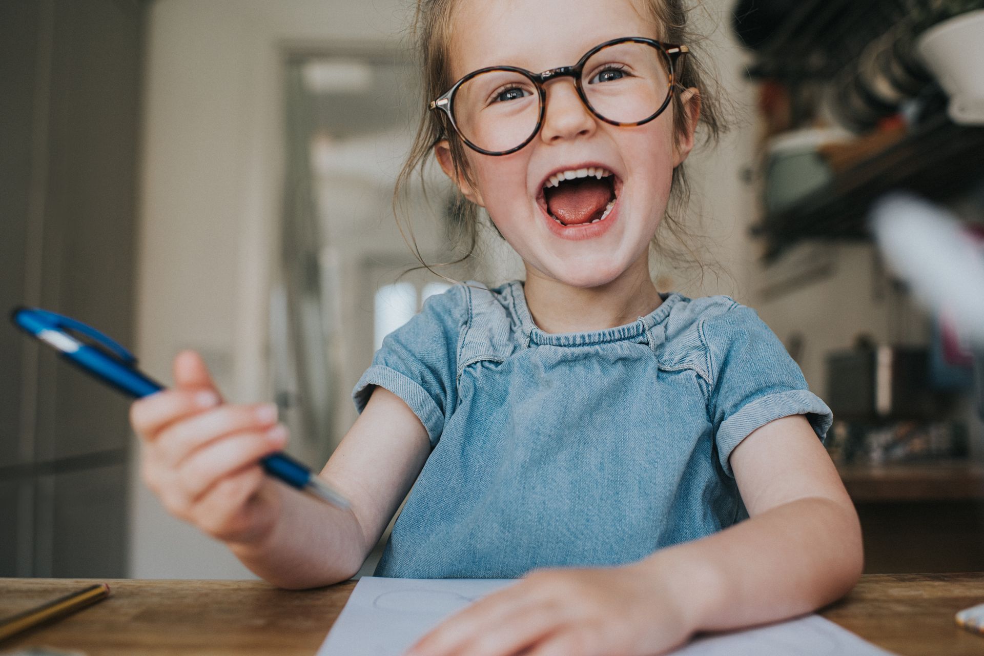 Child with glasses and denim shirt, smiling, holding pen, and sitting at table with paper.