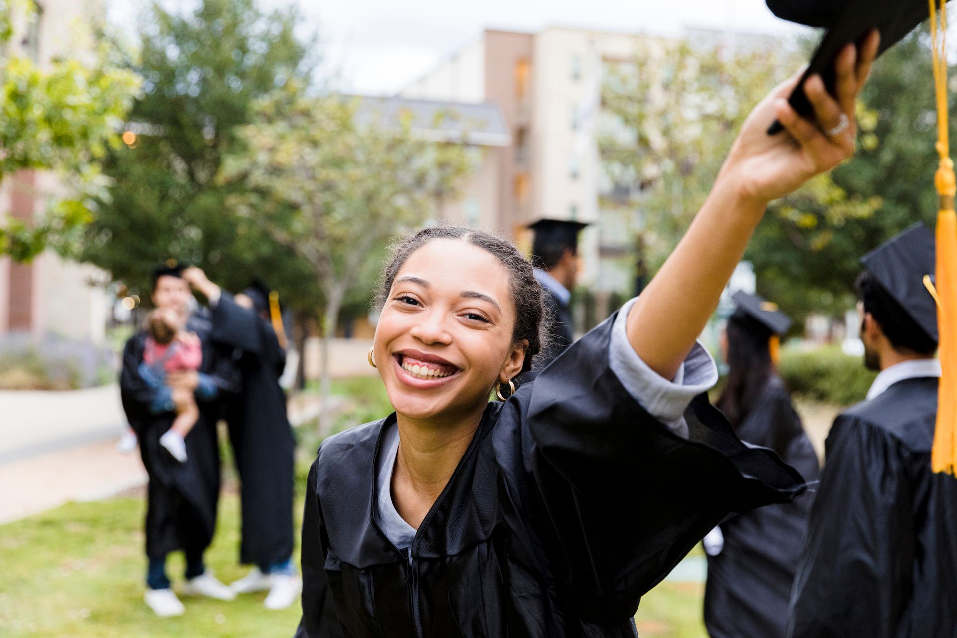 Happy graduate in cap and gown, smiling, raising her cap outside. Other graduates and buildings in background.