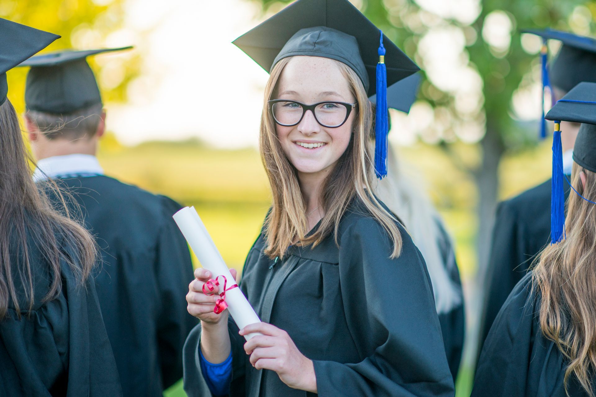 Student in cap and gown smiles, holding diploma, other graduates blurred in background.