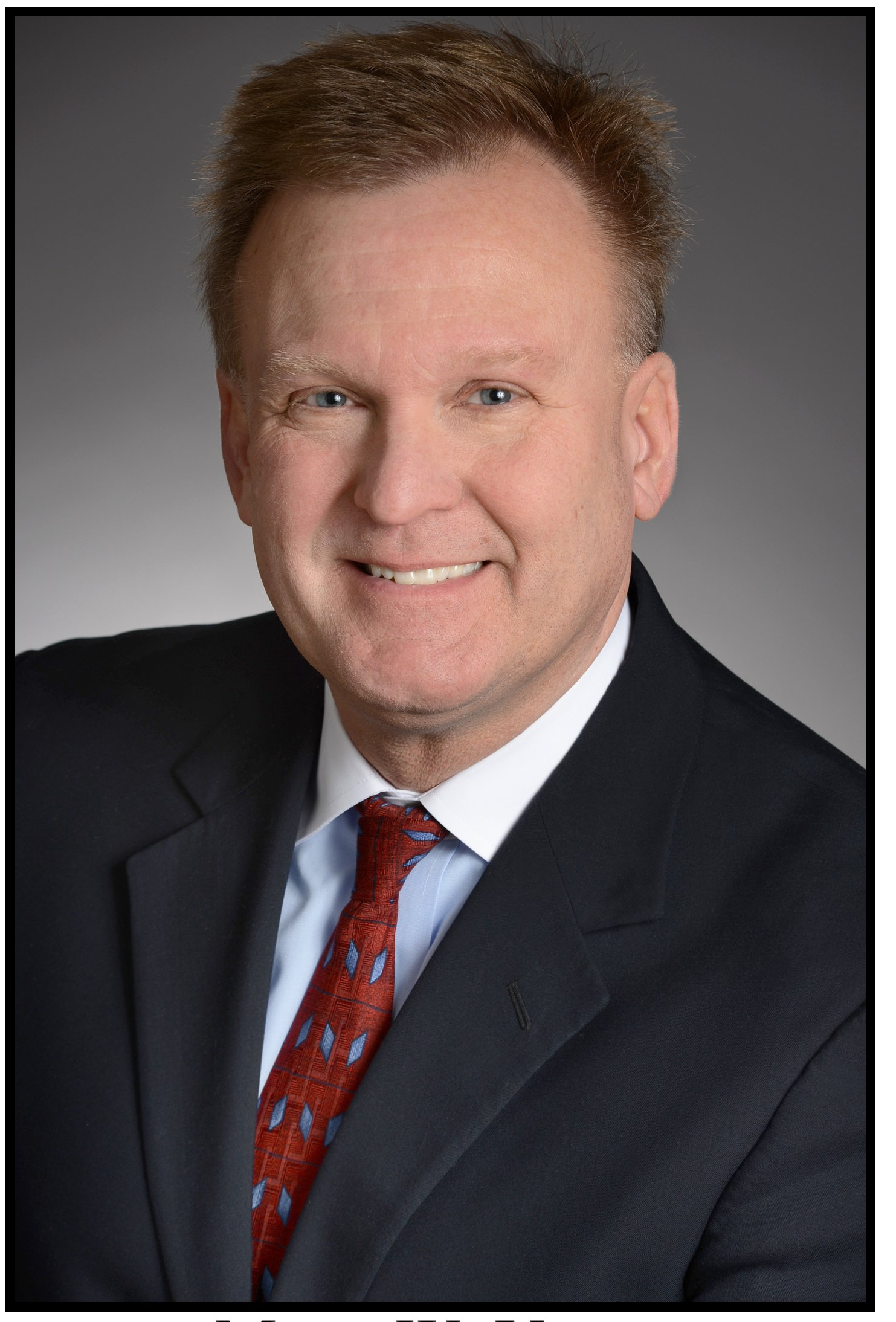 Man in suit smiles at the camera; red patterned tie, blurred background.