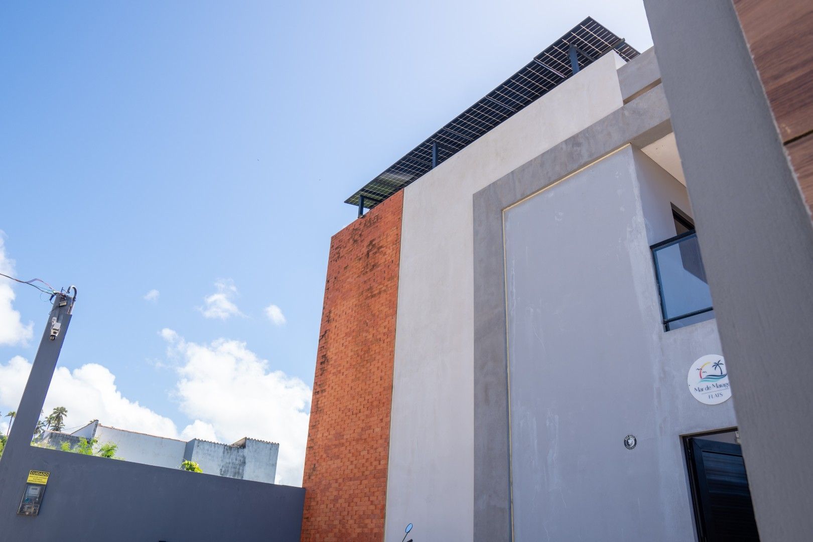 Fachada de edifício moderno com paredes cinzentas, coluna de tijolo e treliça no telhado contra um céu azul.