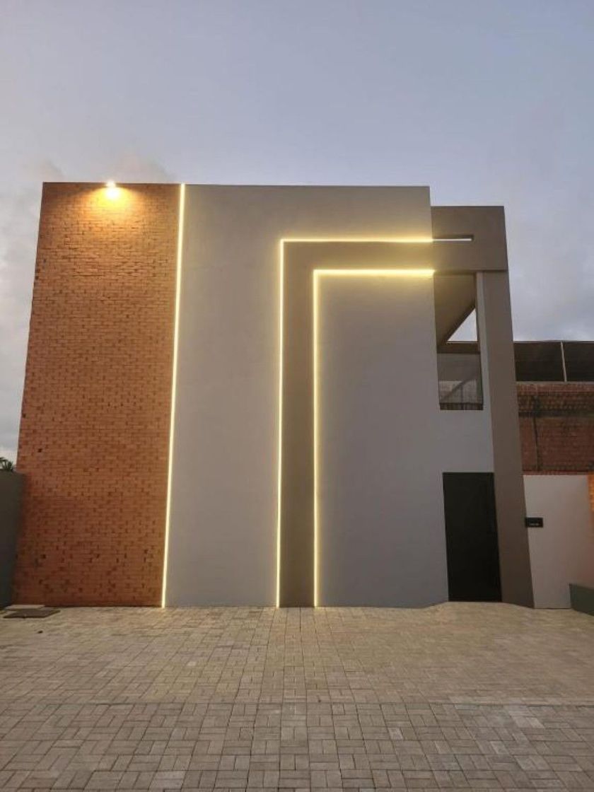 Fachada moderna de edifício com paredes cinzentas, detalhes em tijolo e iluminação LED linear aconchegante ao entardecer.