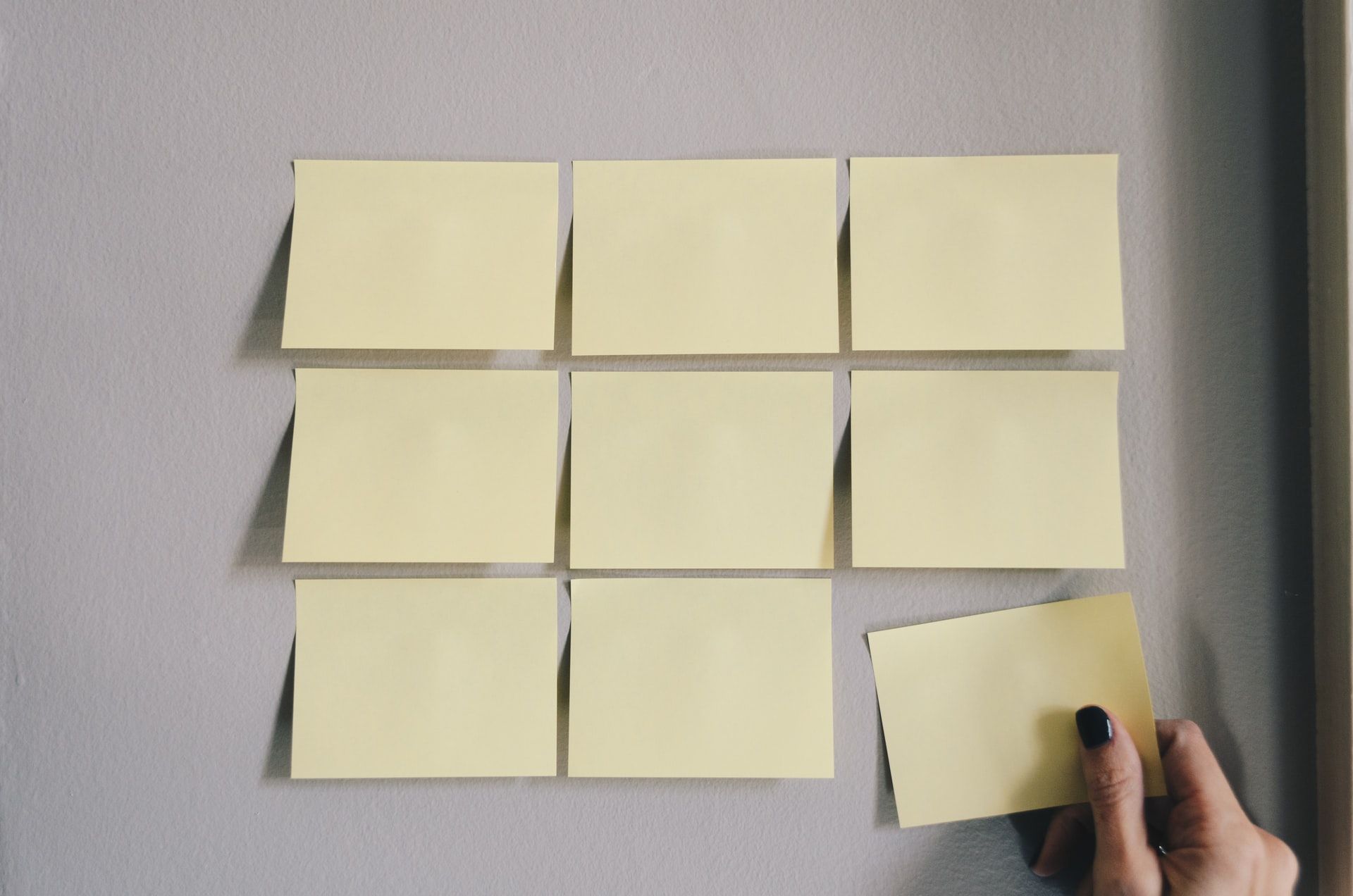 Hand placing a yellow sticky note on a gray wall with eight other blank sticky notes.