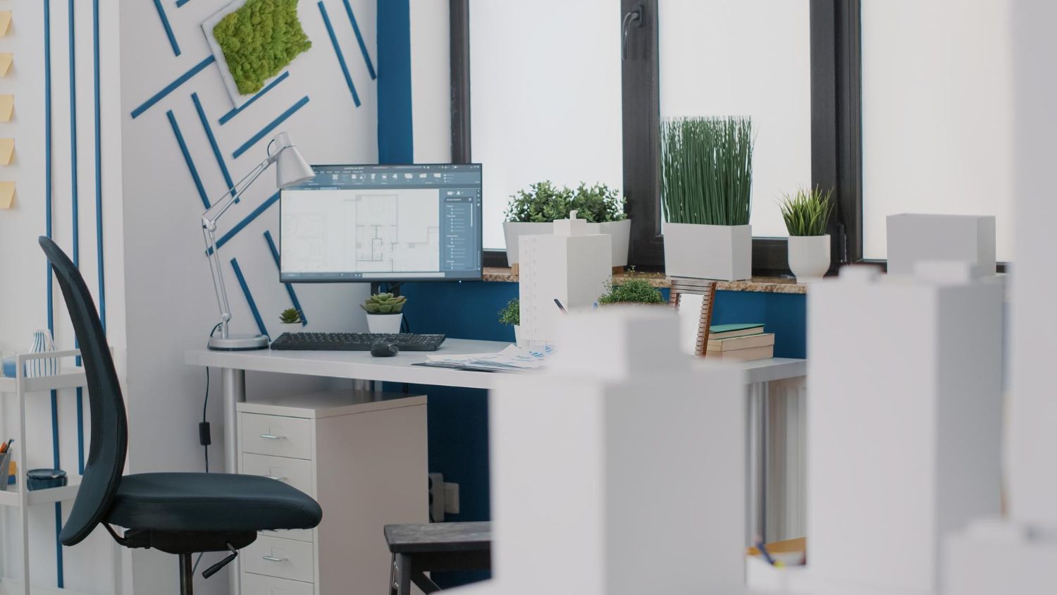 Office desk with computer, window with plants, and decorative white panels.