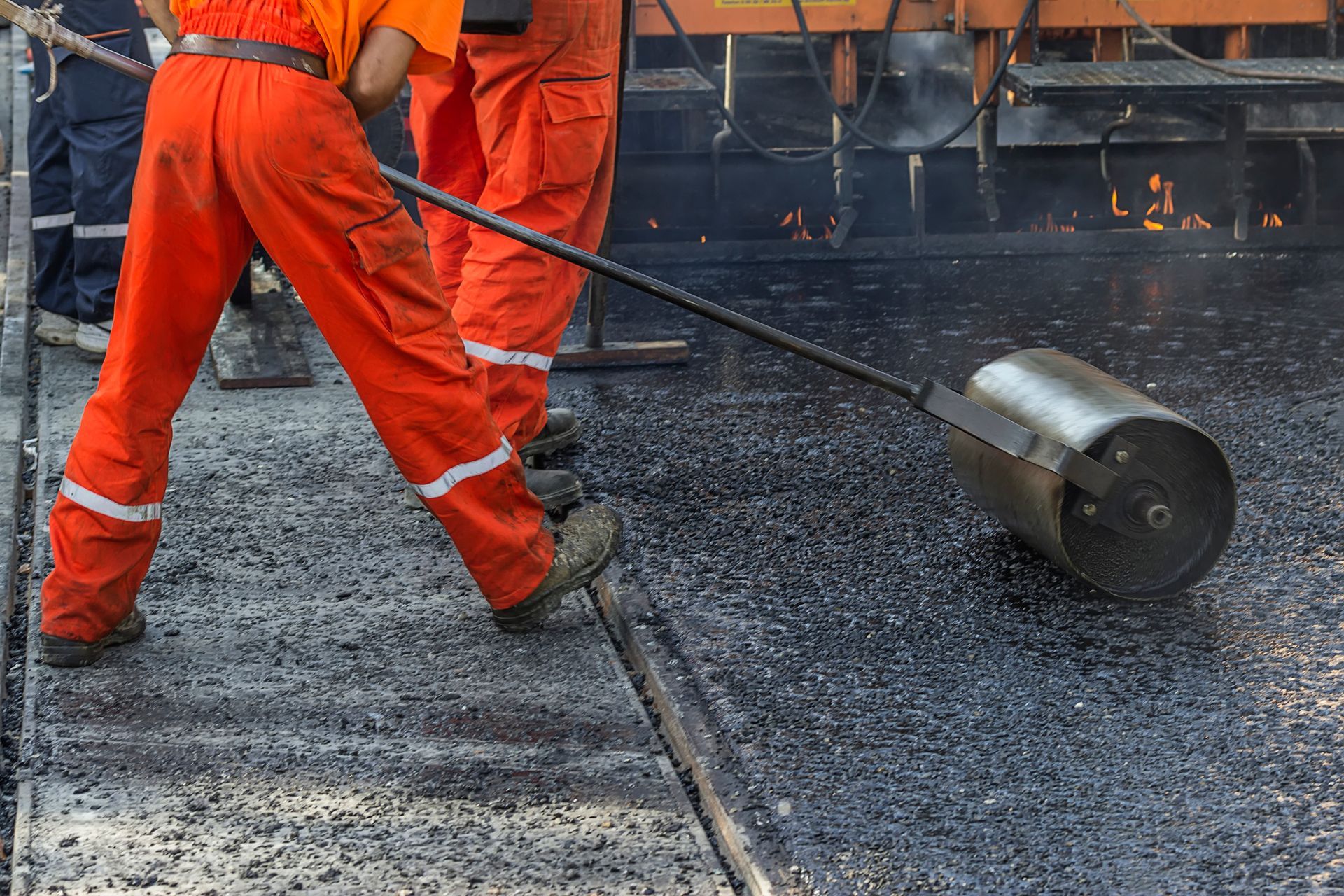 Worker in orange uniform pushes roller over asphalt on the ground, representing paving solutions.
