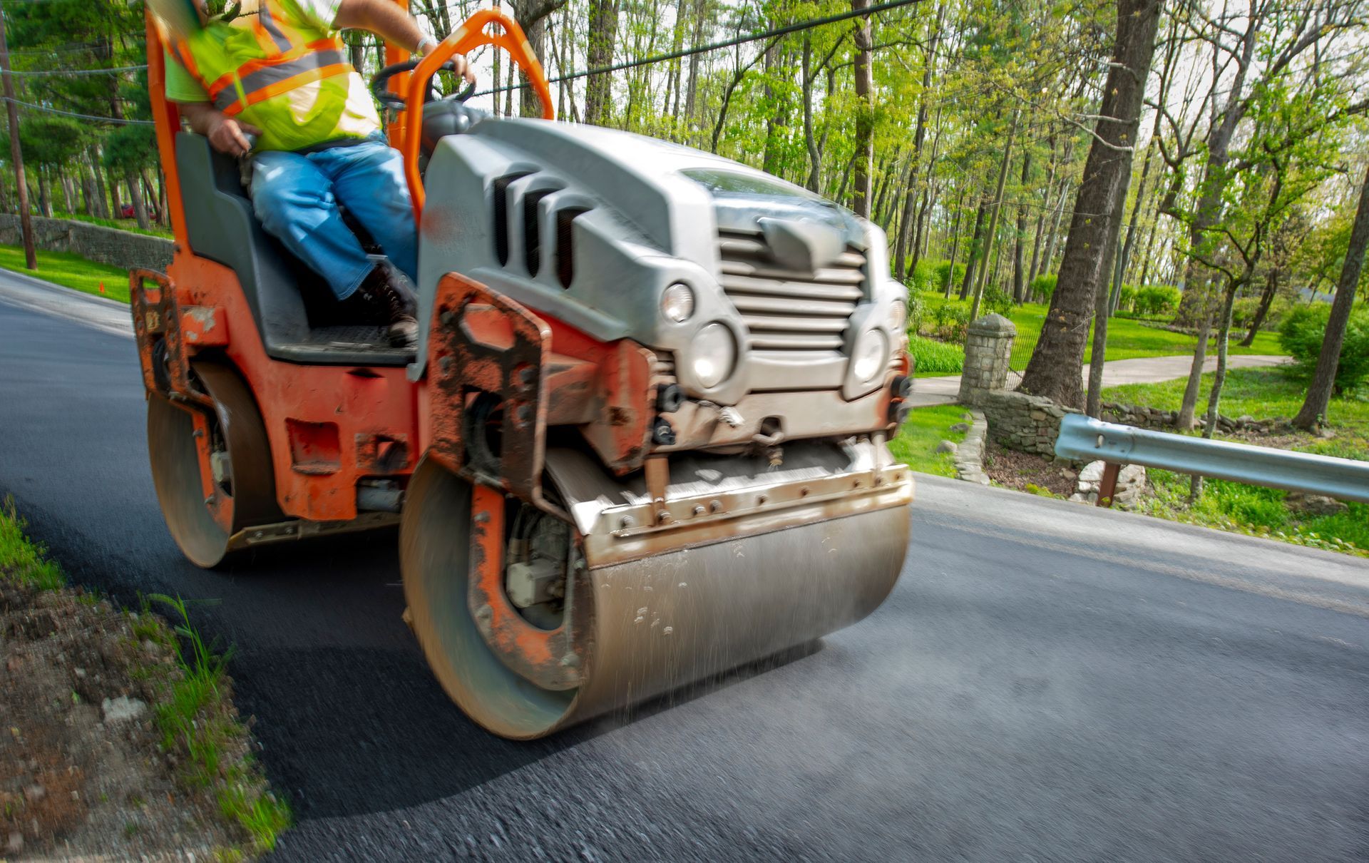 A worker operates a road roller in a wooded area, focusing on asphalt maintenance tasks.