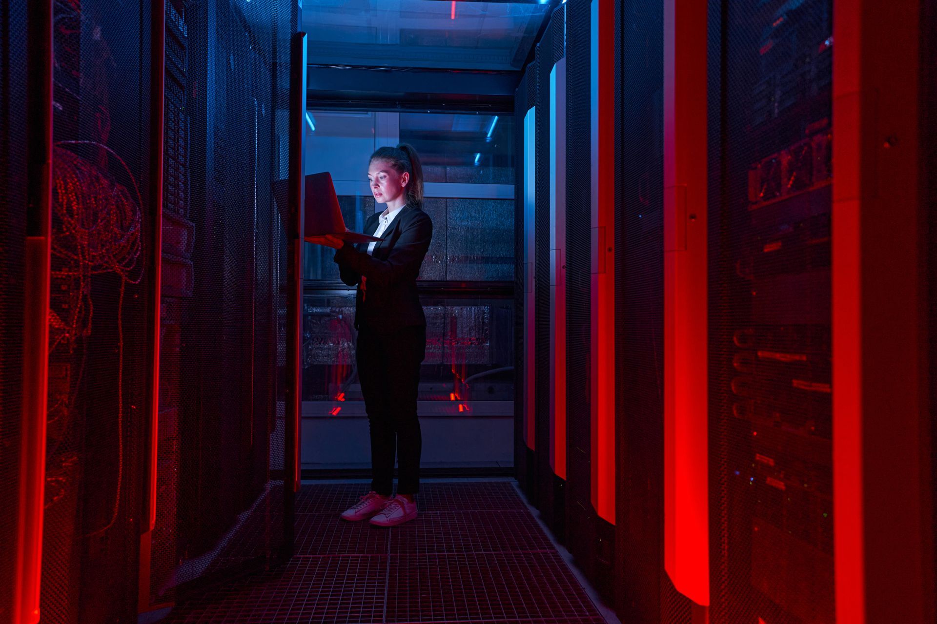 Woman in server room, inspecting equipment with tablet, lit by red and blue lights.