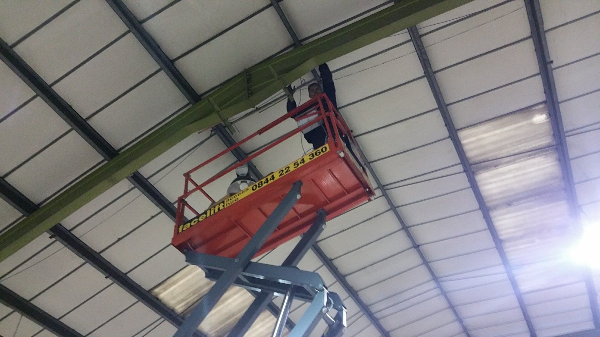 Red maintenance lift raised under a warehouse roof, with a worker on the platform.