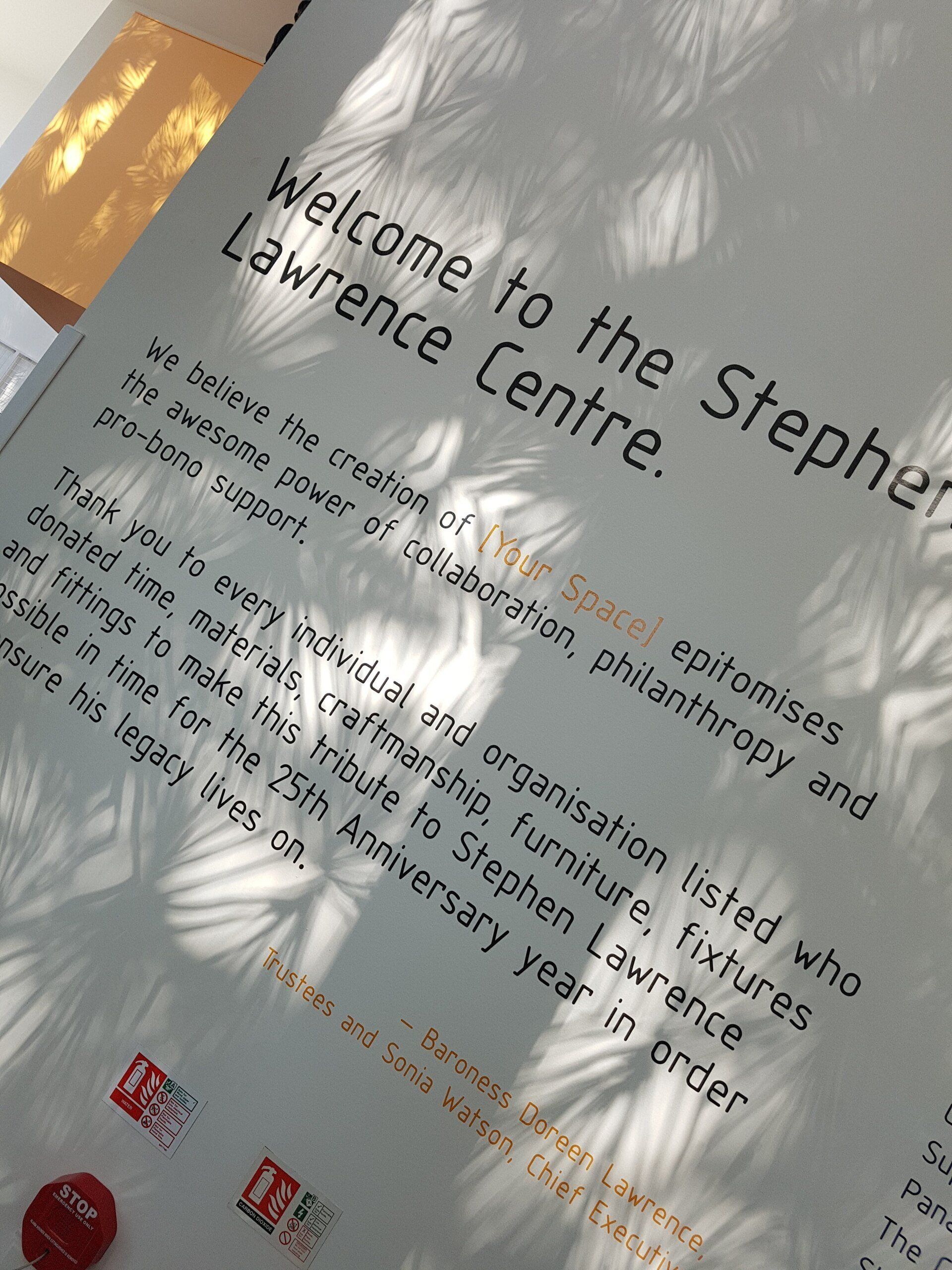 Tilted sign reading “Welcome to the Stephen Lawrence Centre” with sunlit reflections and red logos at the bottom