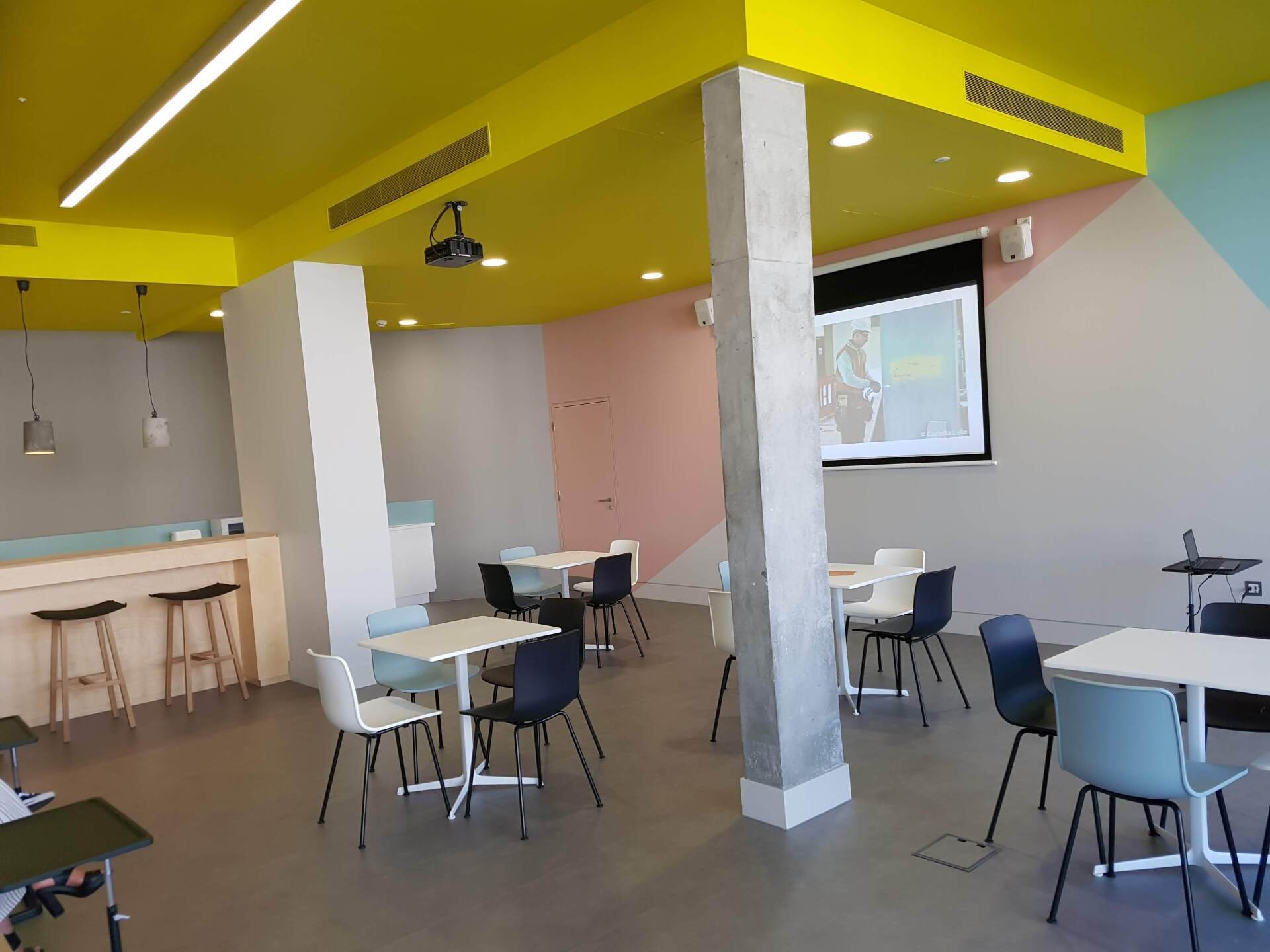 Bright modern café with white tables, chairs, yellow ceiling, and a projection screen on the wall