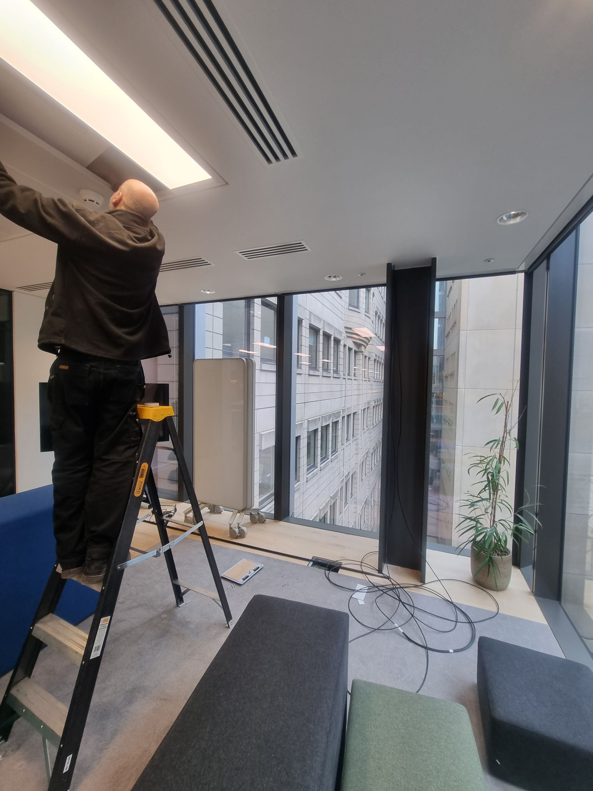 Worker on a ladder fixing a ceiling light in a modern office with city windows.