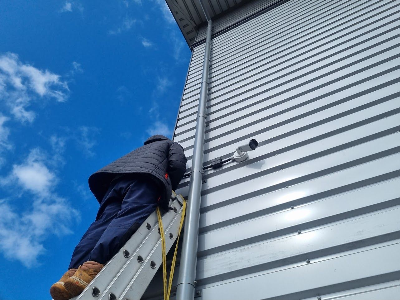 Worker on a ladder cleaning the side of a metal building against a blue sky