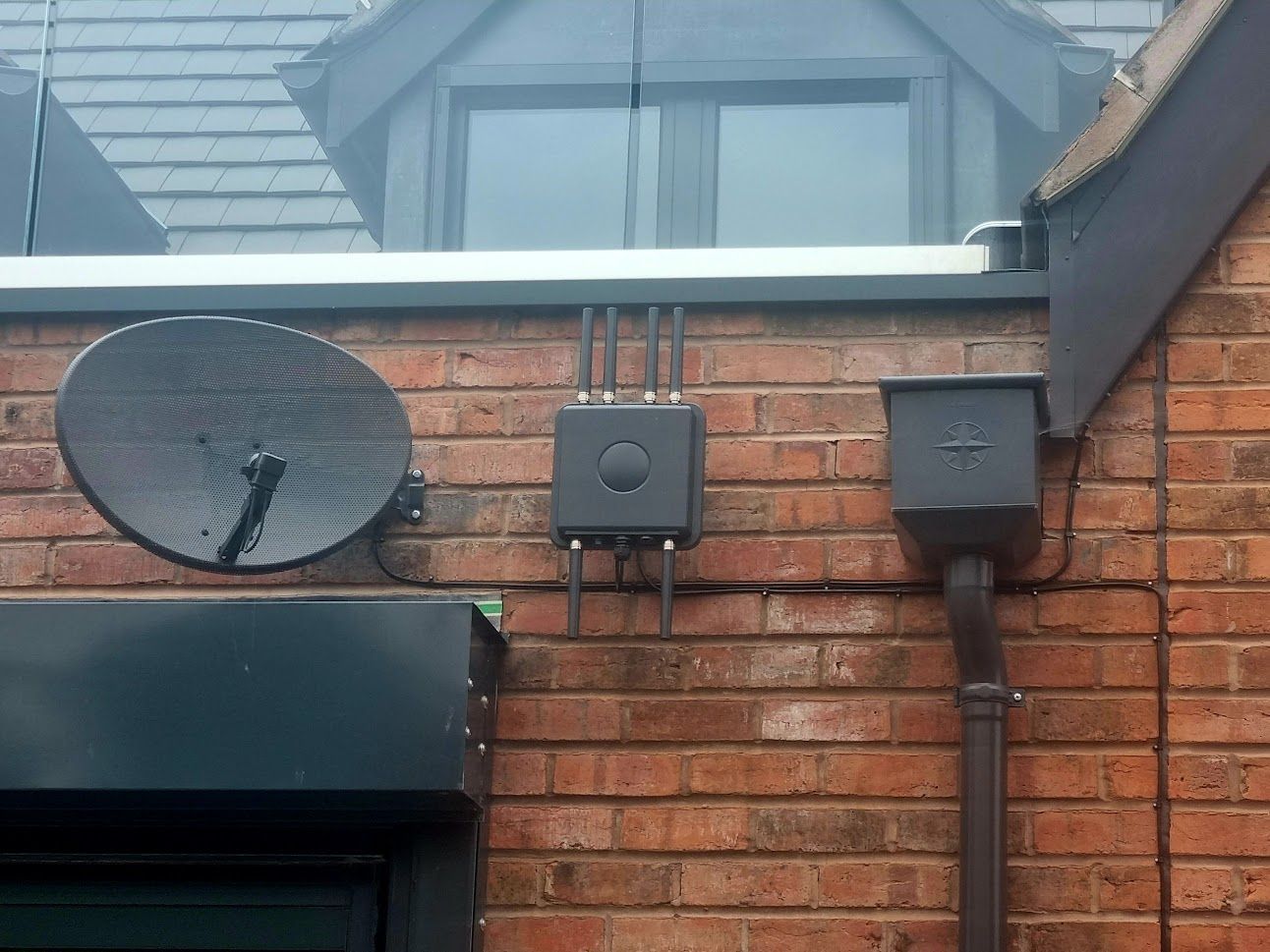 Brick house wall with satellite dish, Outdoor Zyxel Wi-Fi router, and dormer window above a black awning