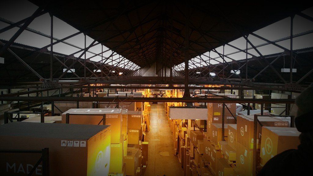 Large indoor warehouse with metal roof trusses and warm yellow lighting over stacked crates and equipment.