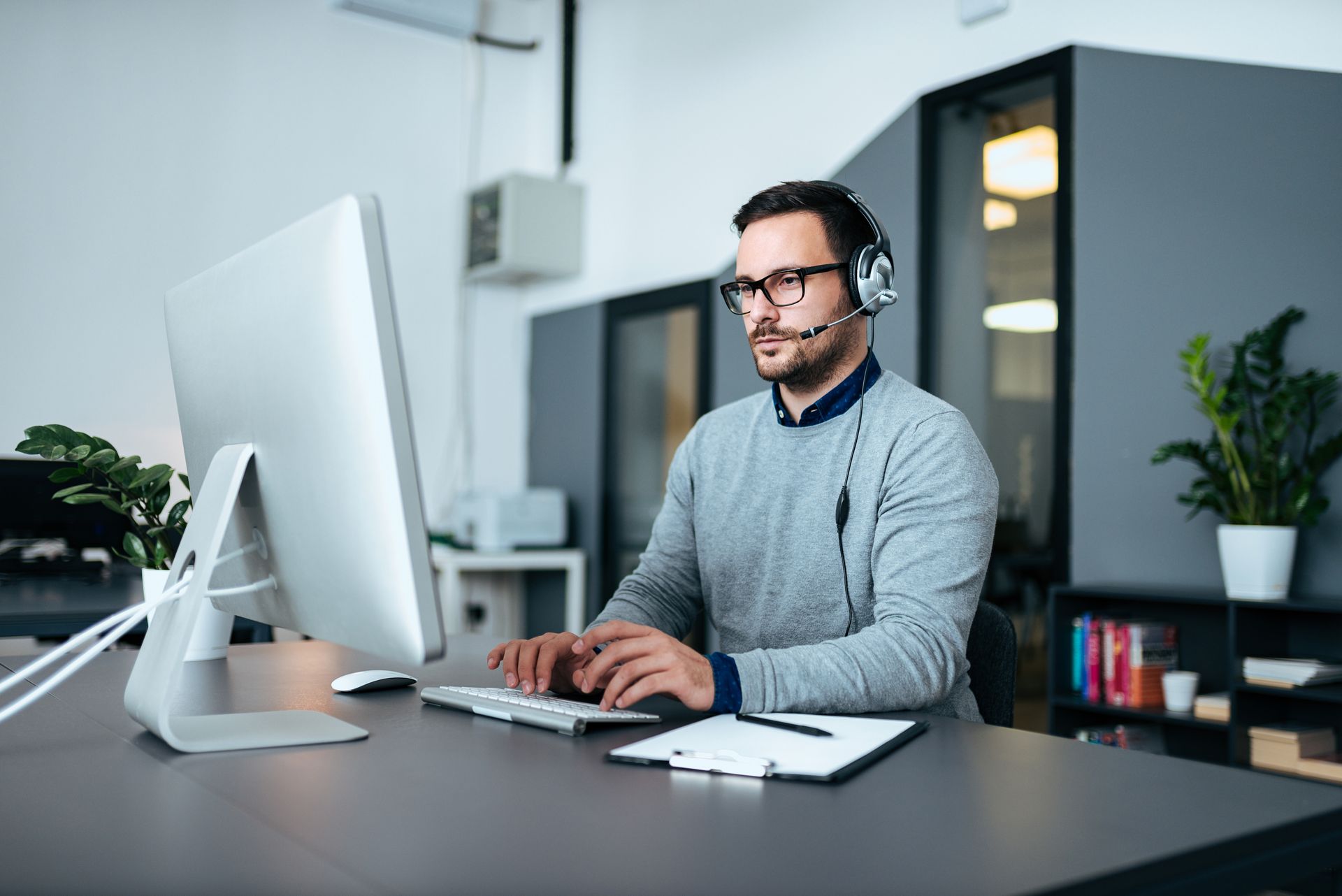Man in glasses and headphones typing at a desktop computer in a modern office