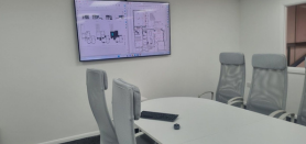 Conference room with white table, gray chairs, and a wall-mounted presentation screen.